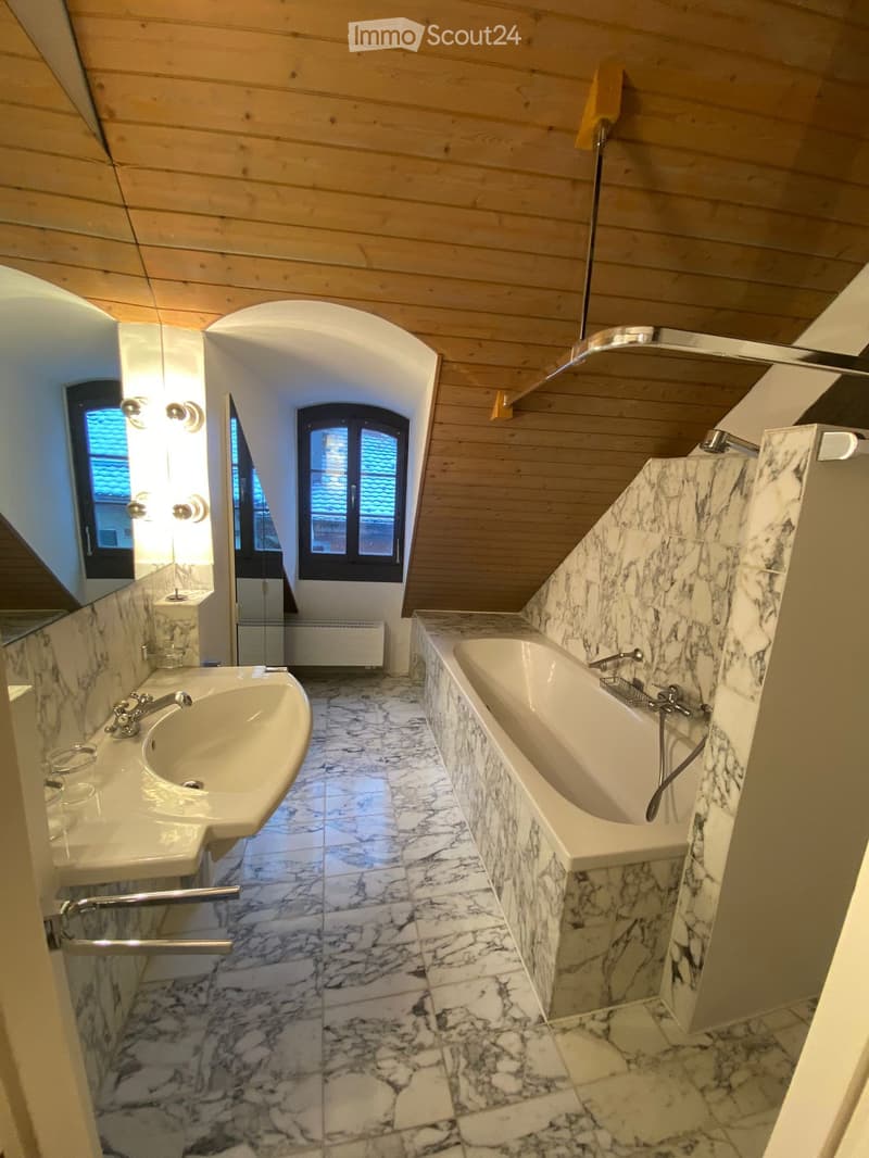 Elegant bathroom with marble tiles, a bathtub, a sink, and a mirror. The room features wooden ceiling beams and arched windows.