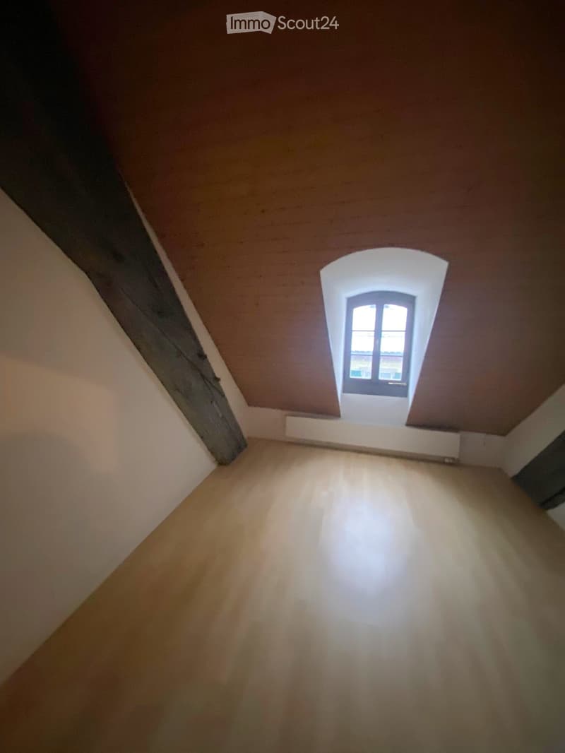 Empty attic space with wooden floor, slanted ceilings, and a small window letting in natural light.