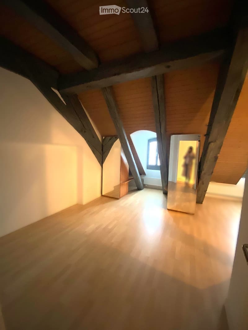 Empty attic space with wooden floors, exposed wooden beams, and a small window letting in natural light.