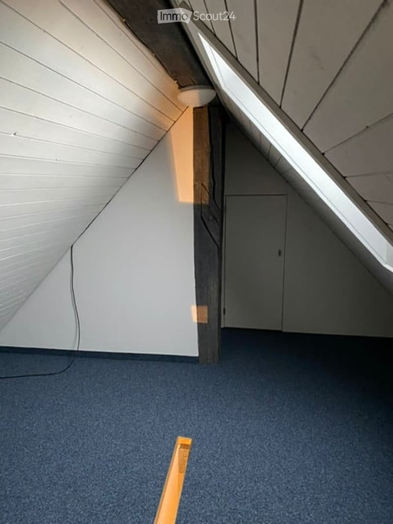An attic space with slanted wooden ceilings, white walls, and a grey closet. The floor is carpeted in blue, and there is a small wooden plank on the floor.