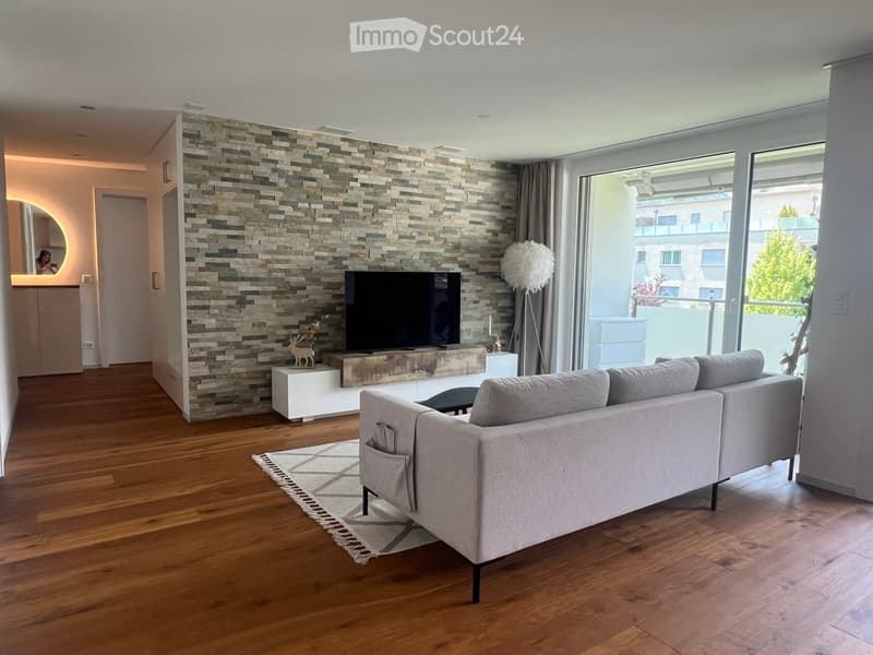 Modern living room with wooden flooring, a grey sectional sofa, a coffee table, a television on a white stand, a floor lamp, and a decorative stone wall.