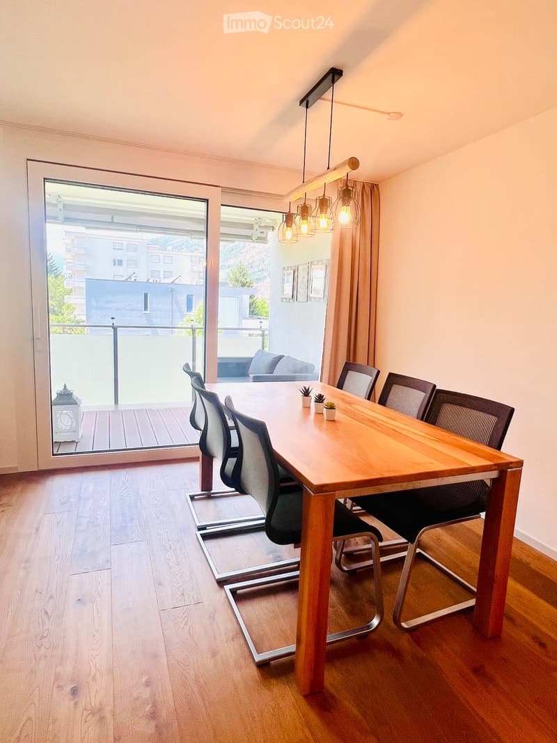 Modern dining area with a long wooden table, several chairs, pendant lights, and a view of a balcony with a sofa and decorative items.