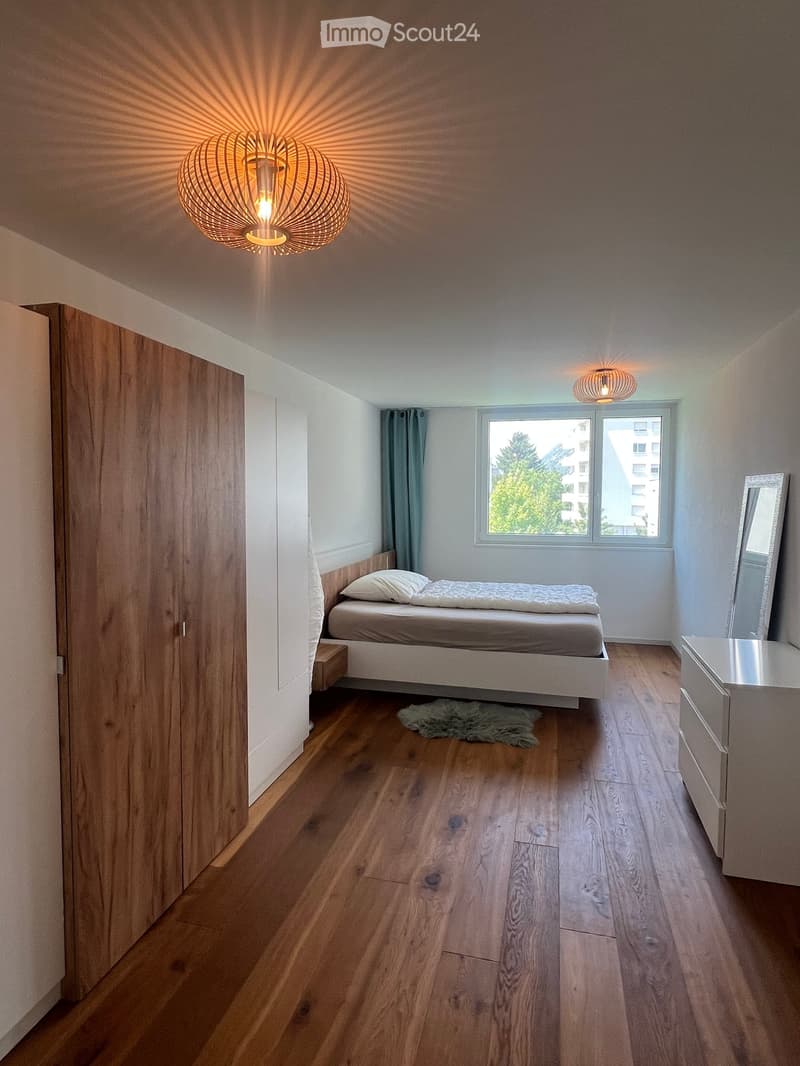 Modern bedroom with wooden flooring, a white bed with pillows and a duvet, a large mirror, a wardrobe, and a nightstand.