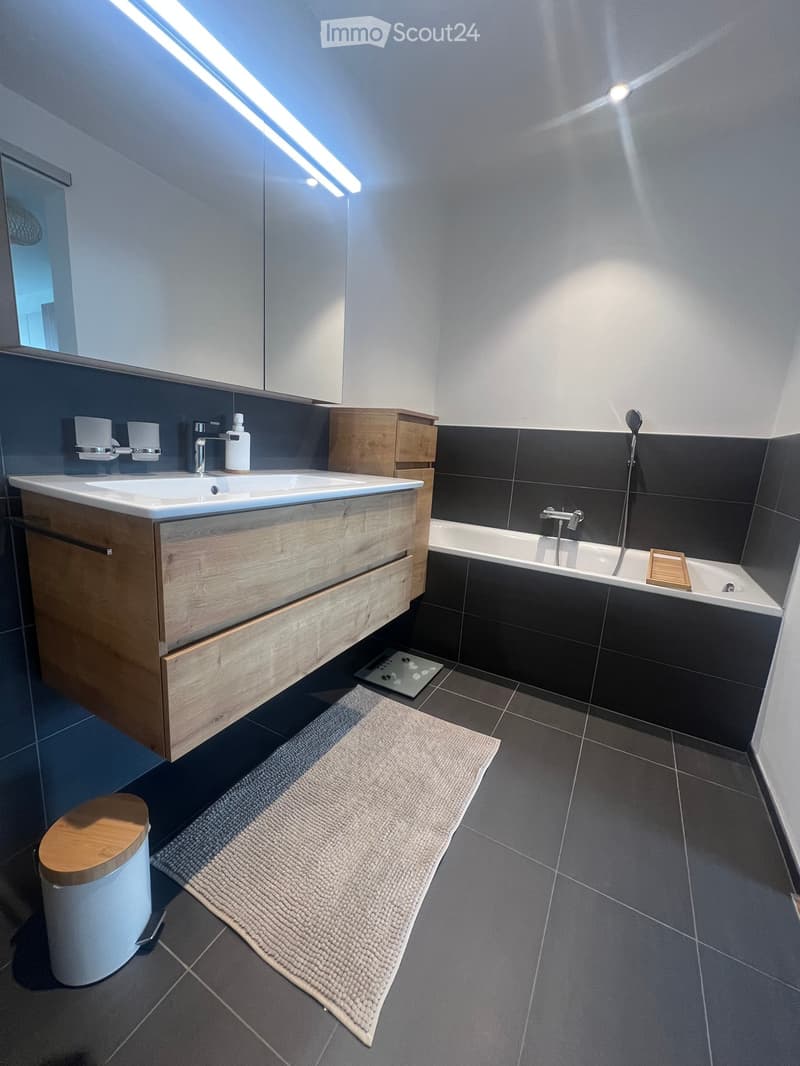 Modern bathroom with a double sink, wooden cabinet, bathtub, and a toilet. Features include a large mirror, sleek fixtures, and a plush rug on the floor.