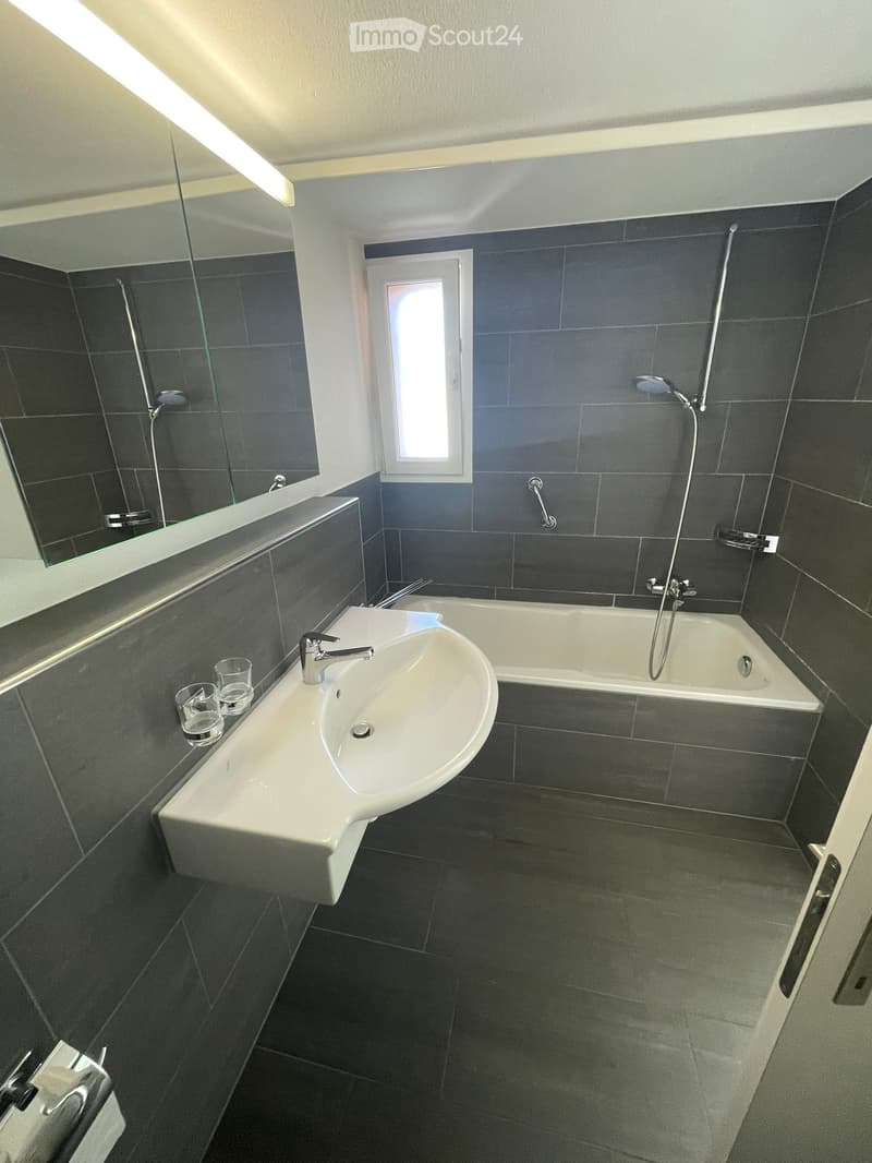 Modern bathroom with dark grey tiles, a white sink, bathtub, and a large mirror. The room features a window and a sleek, contemporary design.