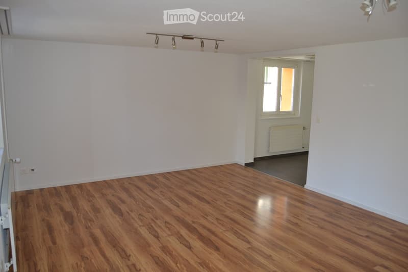 Empty room with wooden flooring and white walls, featuring a track light fixture and a doorway leading to another room with a window and radiator.