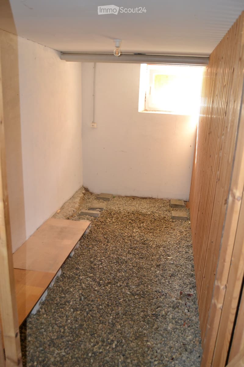 Empty room with a gravel floor, white walls, and a small window letting in natural light. There is a wooden plank on the left side and a few bricks scattered on the floor.