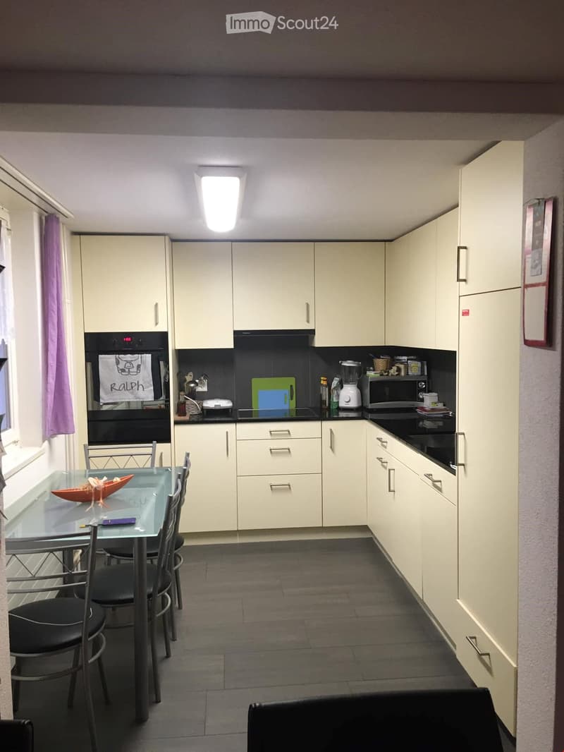 Modern kitchen with white cabinets, black countertops, and a glass dining table with four chairs. Appliances include an oven, microwave, and blender. A decorative towel hangs on the oven door.