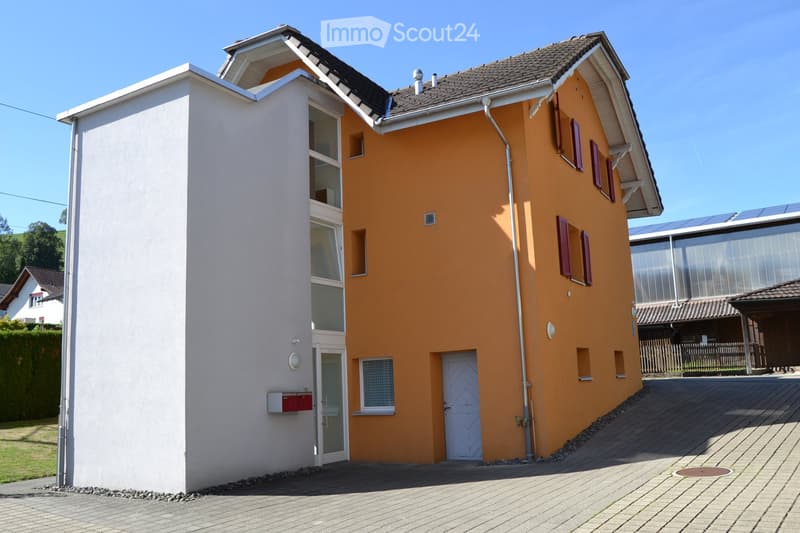 Two-story house with orange and white exterior, multiple windows, and a modern design. Features include a raised ground floor, solar panels on the roof, and a paved driveway.
