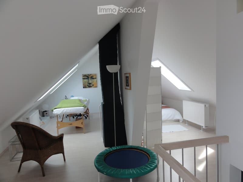 Loft-style room with slanted ceilings, white walls, and wooden flooring. Features a bed, a massage table, a wicker chair, a green exercise ball, a mirror, and a staircase leading to another level.