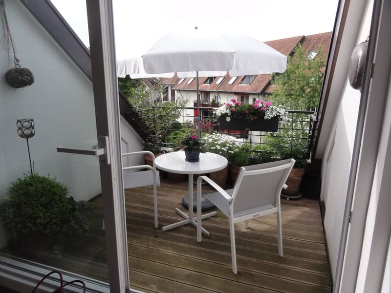Balcony with wooden decking, white table and chairs, umbrella, and potted plants.