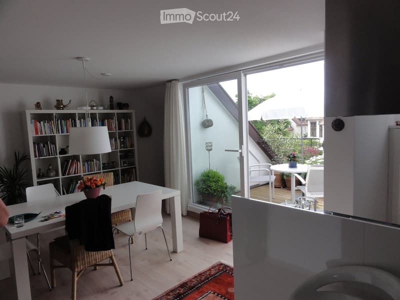 Bright dining area with white table and chairs, bookshelf, pendant lamp, and a view to a balcony with outdoor furniture.