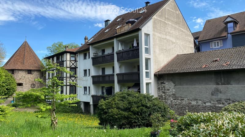 Multi-story residential building with balconies, surrounded by greenery and a stone tower.