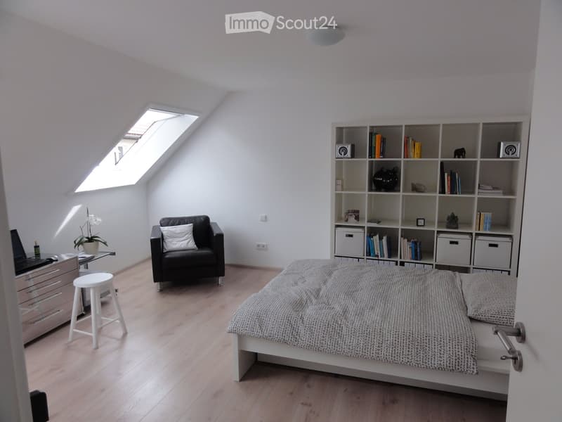 Cozy attic bedroom with a white bed, grey bedding, a black sofa, a small white stool, a laptop on a drawer unit, and a large bookshelf filled with books and decorative items. The room features wooden flooring and a skylight window.