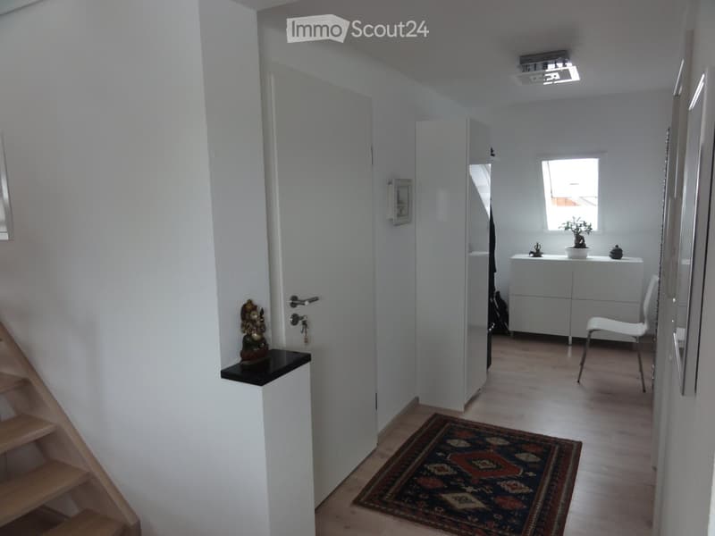 A modern hallway with wooden flooring, a small rug, a white chair, a small table with a plant, and a mirror on the wall.