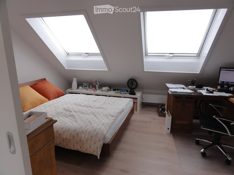 Cozy attic bedroom with slanted ceilings and skylights, featuring a bed with orange pillows, a nightstand, a desk with a computer, and a chair.