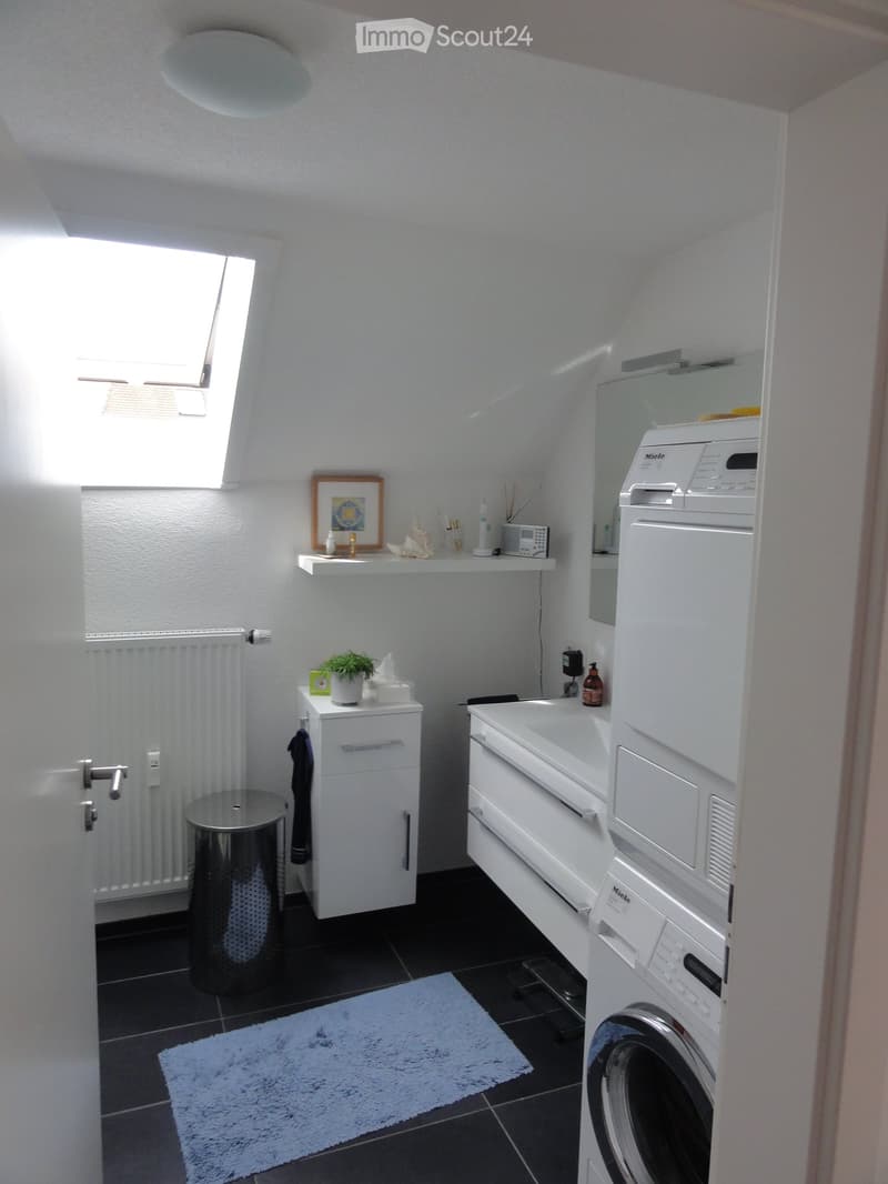 Modern bathroom with white walls, black tiled floor, washing machine, sink, cabinet, mirror, framed picture, plant, and trash can.