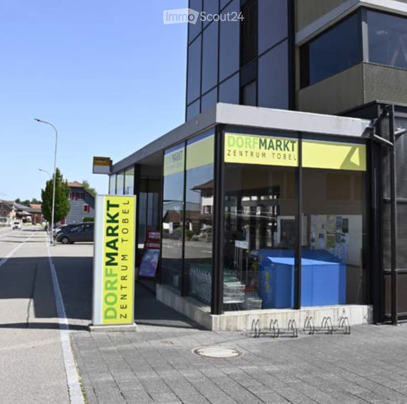 Modern commercial building with large glass windows and signage reading "Dorfmarkt Zentrum Tobel".