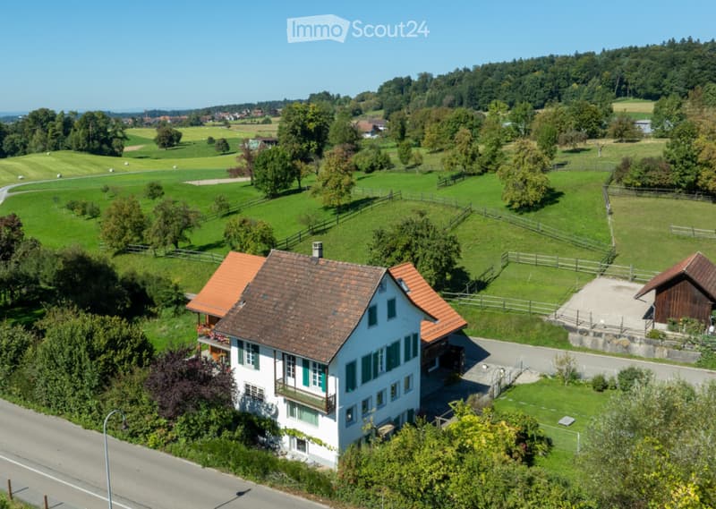 A two-story white house with a red-tiled roof, surrounded by green fields and trees. The house features multiple windows and a small balcony on the upper floor. A wooden outbuilding is visible to the right, and the property is enclosed by a fence.