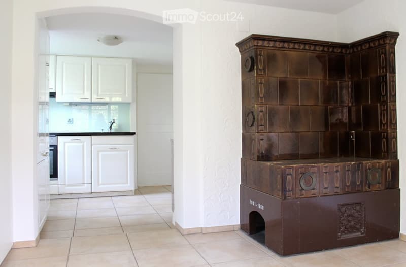 A modern kitchen with white cabinets, a sink, and a wooden cabinet with intricate carvings. The floor is tiled, and the walls are white.