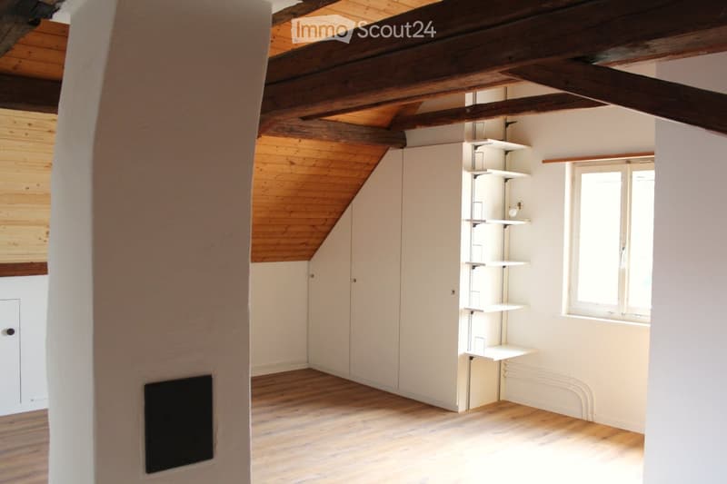 Empty attic room with wooden beams, white walls, wooden floor, built-in shelving unit, and a window letting in natural light.