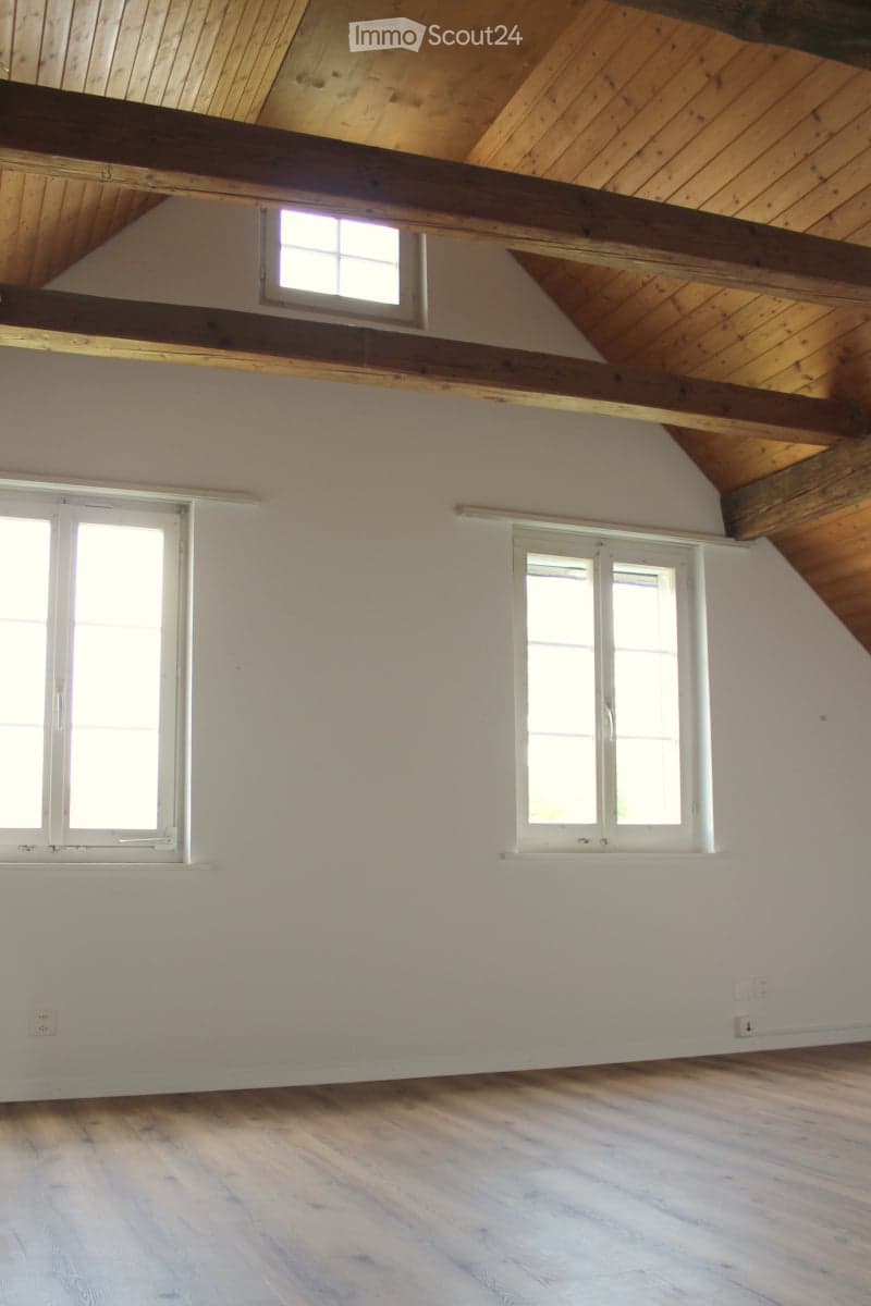 Empty room with wooden flooring, white walls, and exposed wooden beams on the ceiling. Three windows provide natural light.