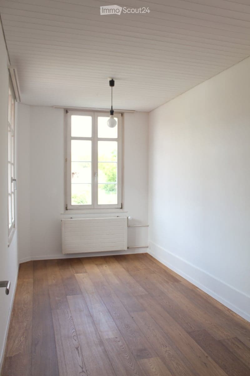 Empty room with wooden flooring, white walls, a window with white frame, a radiator, and a hanging light fixture.