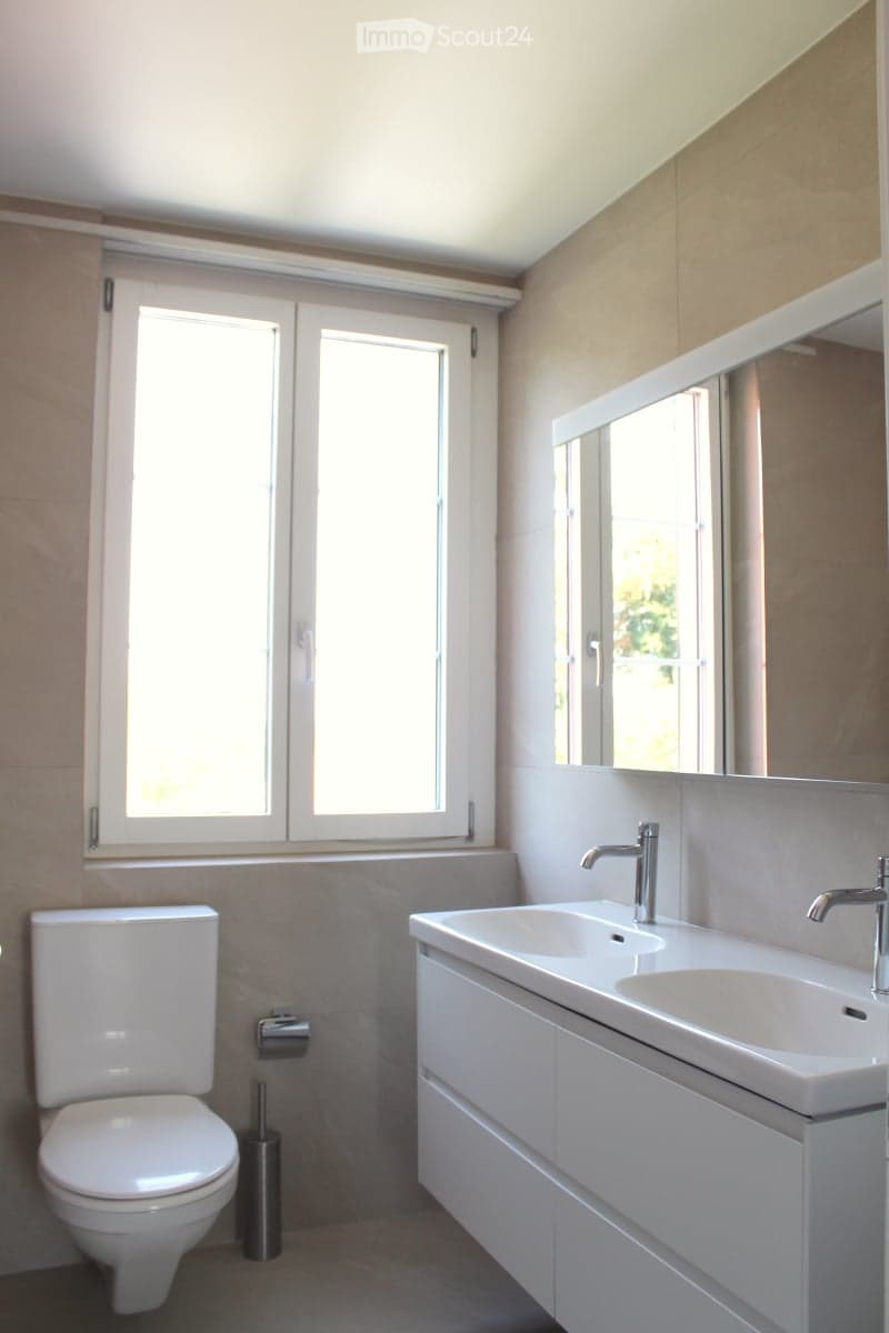 Modern bathroom with a white toilet, double sink, mirrored cabinet, and large windows allowing natural light.
