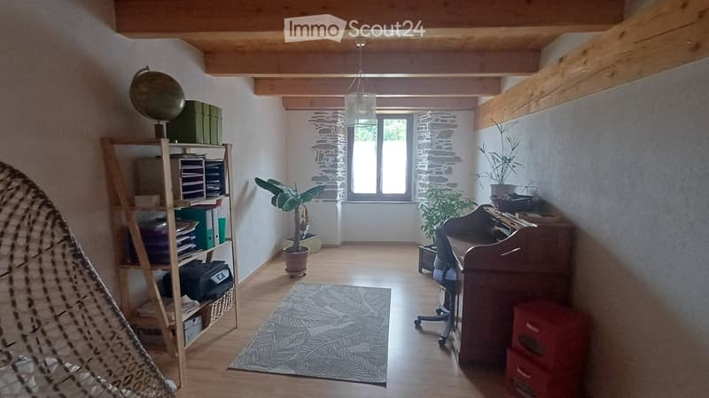 A cozy, well-lit room with wooden floors, exposed wooden beams, and a large window. Features include a wooden desk with a chair, a shelf with books and storage boxes, a hanging globe, a potted plant, and a patterned rug.