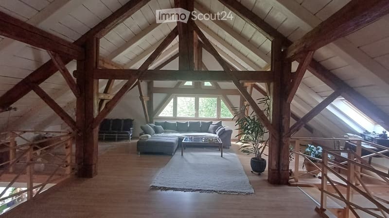 Spacious living room with exposed wooden beams, large windows, grey sectional sofa, coffee table, ottoman, and potted plants.