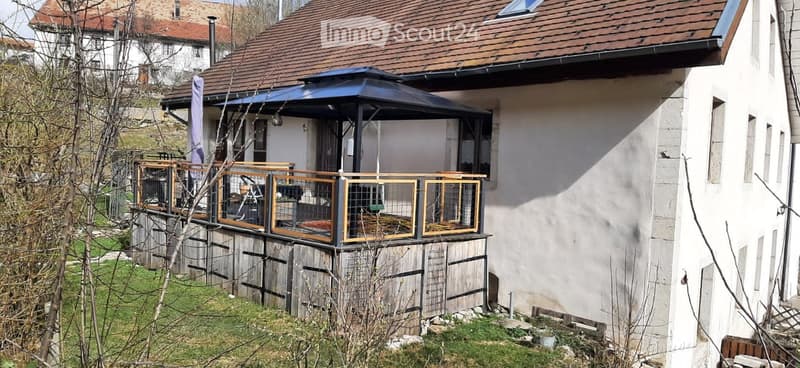 Balcony with wooden railing and a covered area, attached to a house with a tiled roof and white exterior walls.