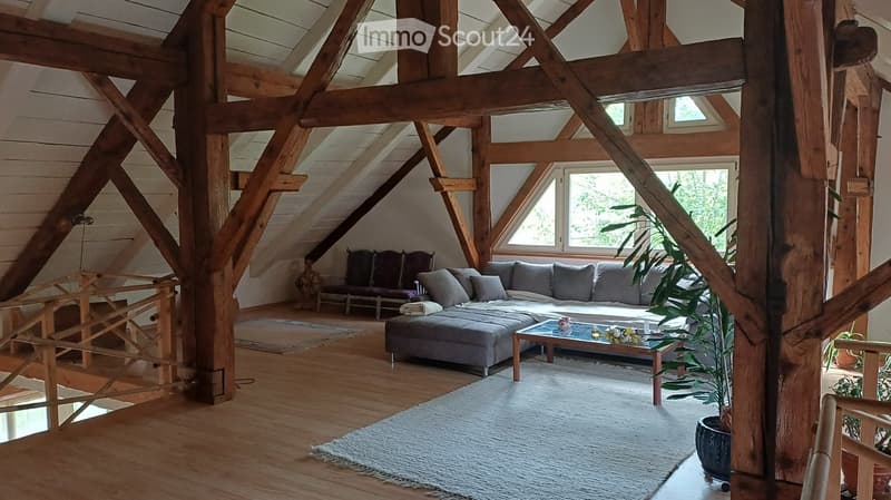 Spacious living room with wooden beams, large windows, grey sectional sofa, coffee table, ottoman, and potted plants.