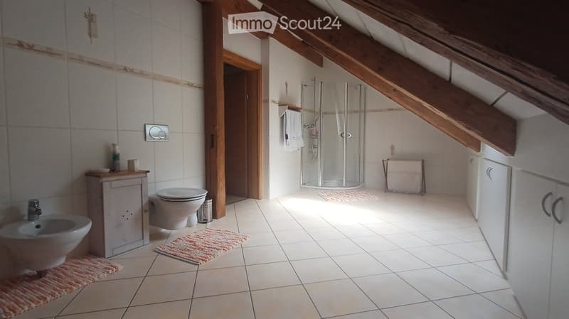 Bright bathroom with white tiles, exposed wooden beams, a toilet, bidet, shower, and a small cabinet with a bottle on top.