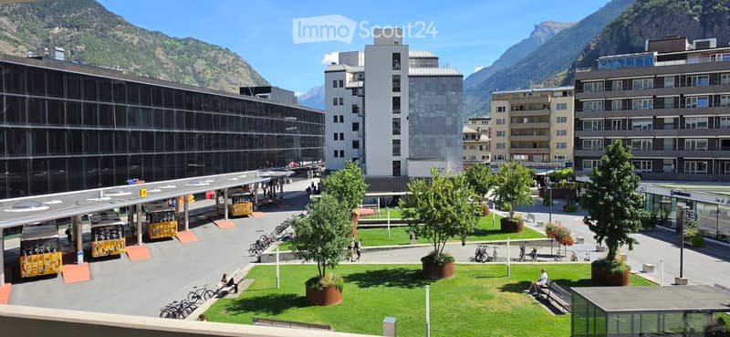 A modern urban square with a mix of modern and traditional buildings, surrounded by mountains. The area features a paved walkway, benches, trees in planters, and a few people walking or sitting. There are bicycles and a bus stop with a yellow bus visible.