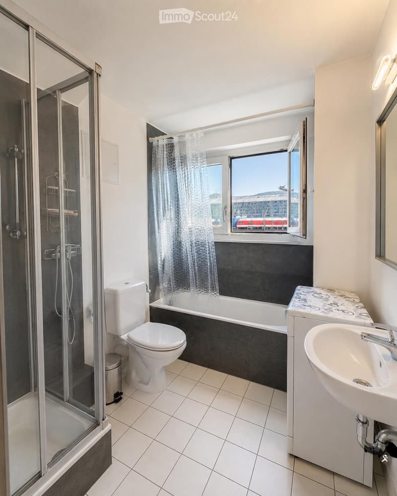 Modern bathroom with white tiles, a bathtub, toilet, sink, and a glass-enclosed shower. A large window offers a view of the city.