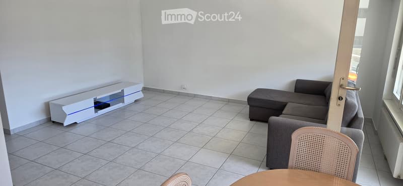Modern living room with tiled floor, grey sectional sofa, wooden coffee table, wicker chair, and white TV stand with blue lighting.