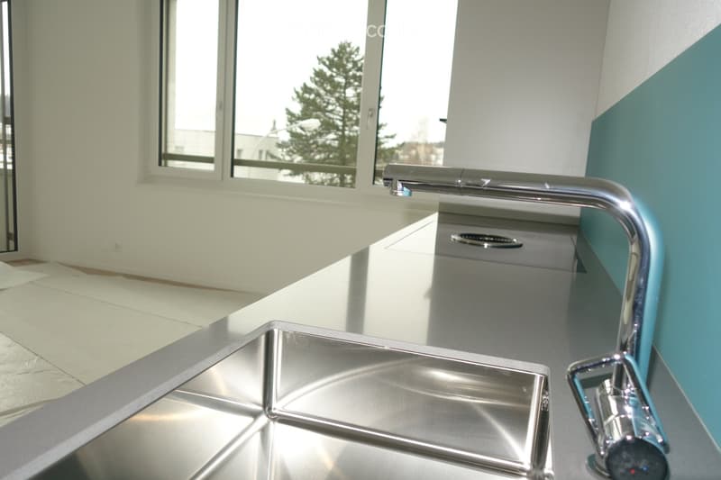 Modern kitchen with stainless steel sink and faucet, white countertops, and a large window overlooking a green area.