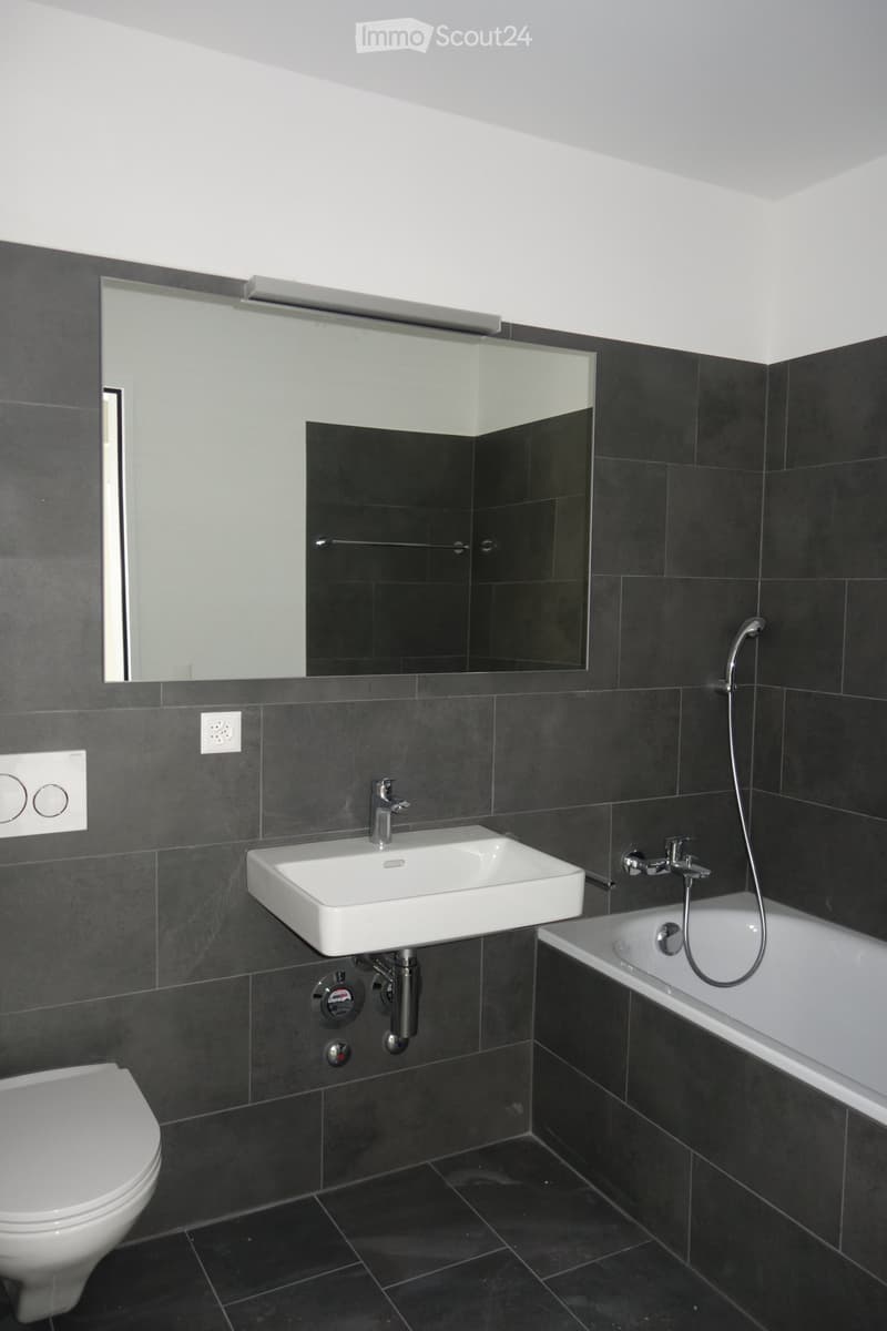 Modern bathroom with dark tiled walls and floor, white sink, bathtub, toilet, and large mirror above the sink.