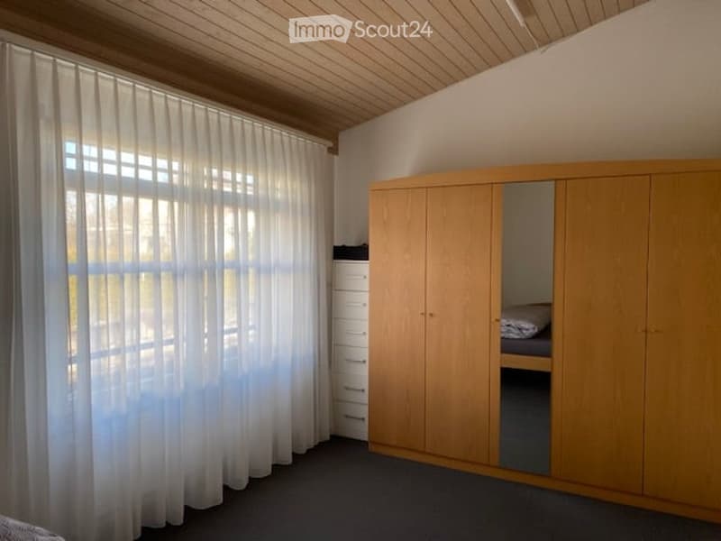 The room features a large wooden wardrobe with mirrored doors, a drawer unit, and a bed tucked inside the wardrobe. Natural light filters through sheer white curtains covering a large window. The ceiling has a wooden panel design.