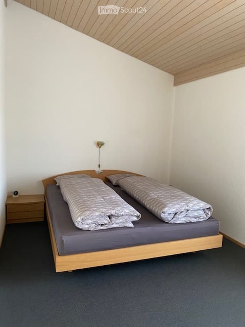 A small bedroom with a single bed, two pillows, and a nightstand with an alarm clock. The room has white walls and a wooden ceiling.