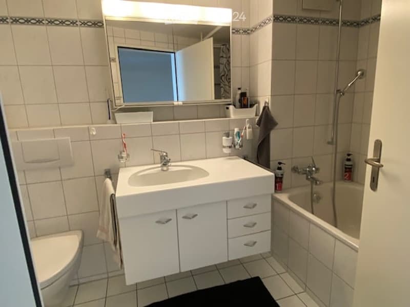 Modern bathroom with white tiles, a sink with a mirror above it, a bathtub, and a toilet. The sink has a modern faucet and a cabinet below. A towel hangs on the wall, and various toiletries are visible on the sink counter.