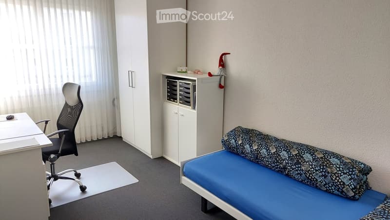A small, furnished bedroom with a single bed, desk, chair, and cabinet. The room features a grey carpeted floor, white walls, and a window with sheer curtains.