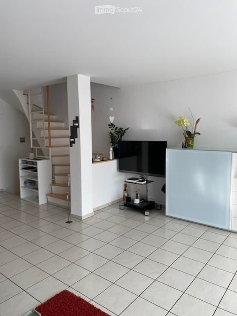 A modern living room with a TV on a stand, a small cabinet, a shelf, and a plant on the windowsill. The space features tiled flooring and a staircase leading upwards.