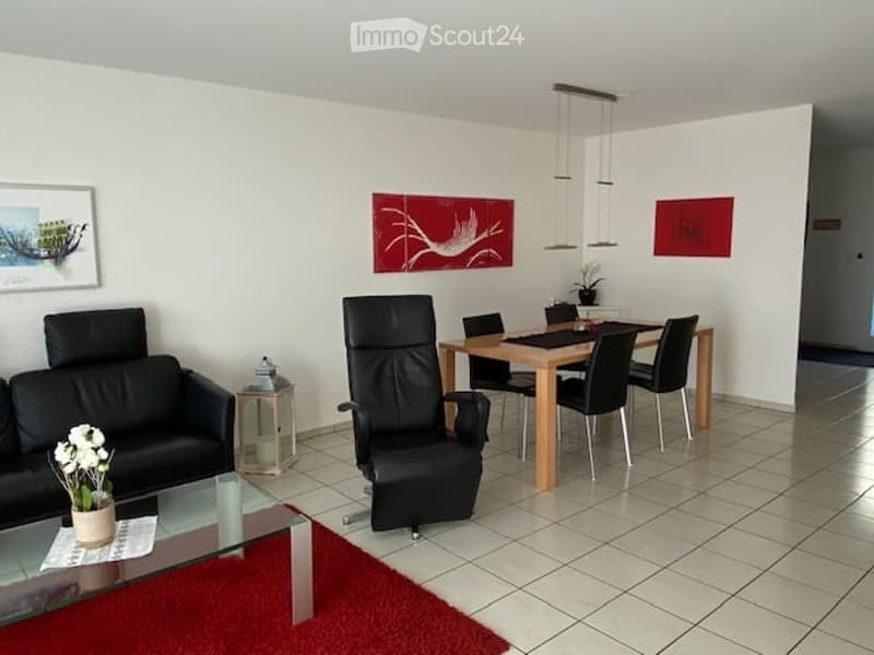 Modern open-plan living and dining area with white walls, tiled flooring, black leather sofa, black leather chair, wooden dining table with black chairs, side table with a flower vase, and artwork on the walls.