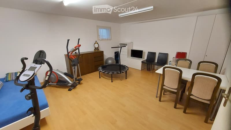 A well-lit room with wooden flooring, featuring exercise equipment like a treadmill and exercise bike, a small table with chairs, a screen, and a clock on a cabinet.