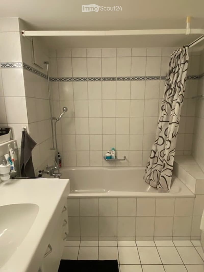 Clean, modern bathroom with white tiled walls and floor, a bathtub with a shower curtain, a sink with a faucet, and a towel hanging on the side.