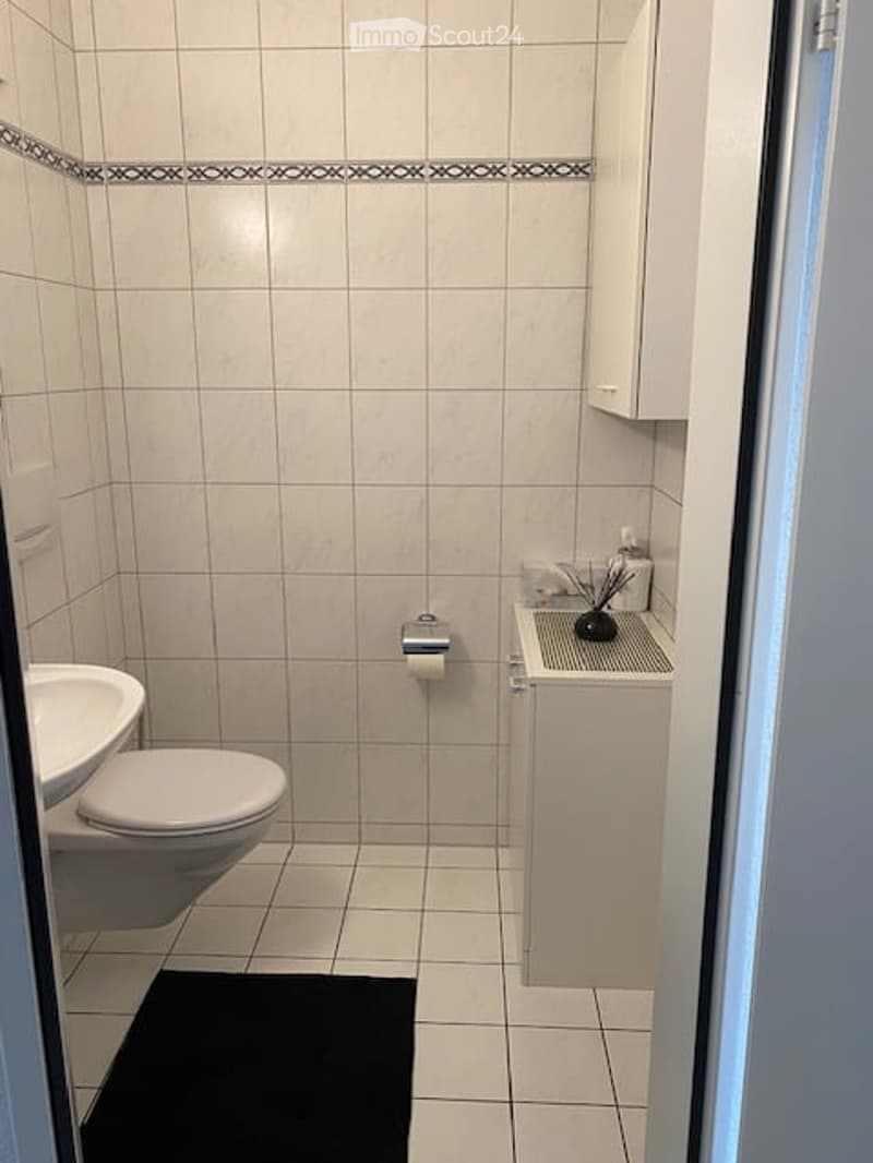 Small bathroom with white tiled walls and floor, toilet, sink, cabinet, and a black rug on the floor.