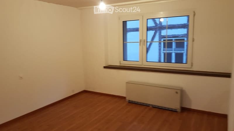 Empty room with wooden flooring, white walls, and a radiator under a window. No furniture or personal items are present.