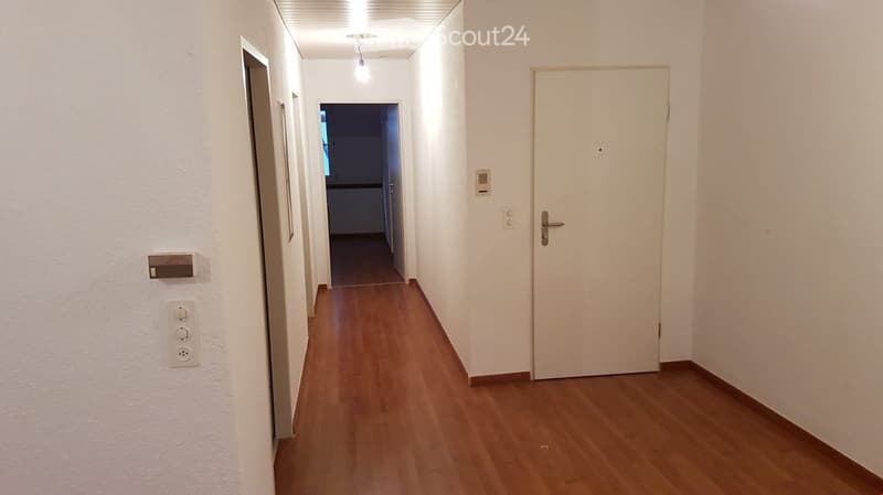 A narrow hallway with wooden flooring, white walls, and a closed door at the end. The hallway features light fixtures and electrical outlets on the walls.