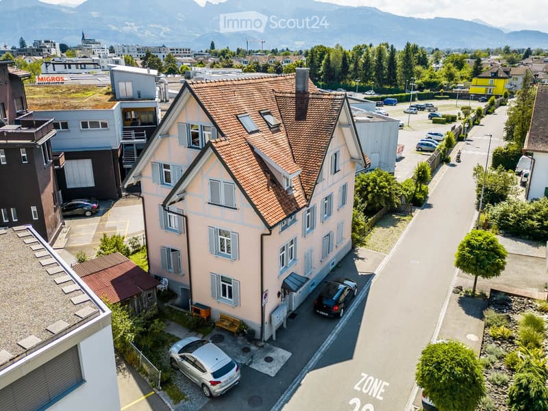 pink exterior, brown roof, multiple windows, car parked in front, located in front of other buildings with mountain views